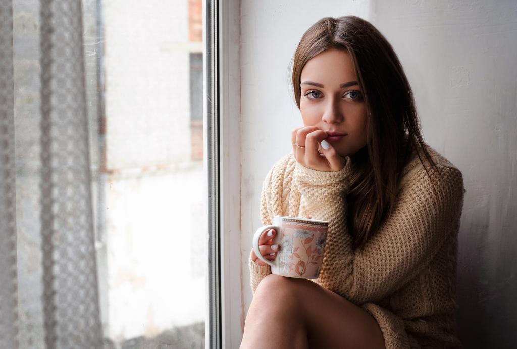 一件毛衣的年轻女孩坐在窗台上的一杯