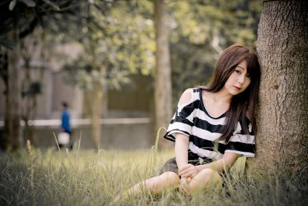 年轻的亚洲女孩坐在树附近的草地上