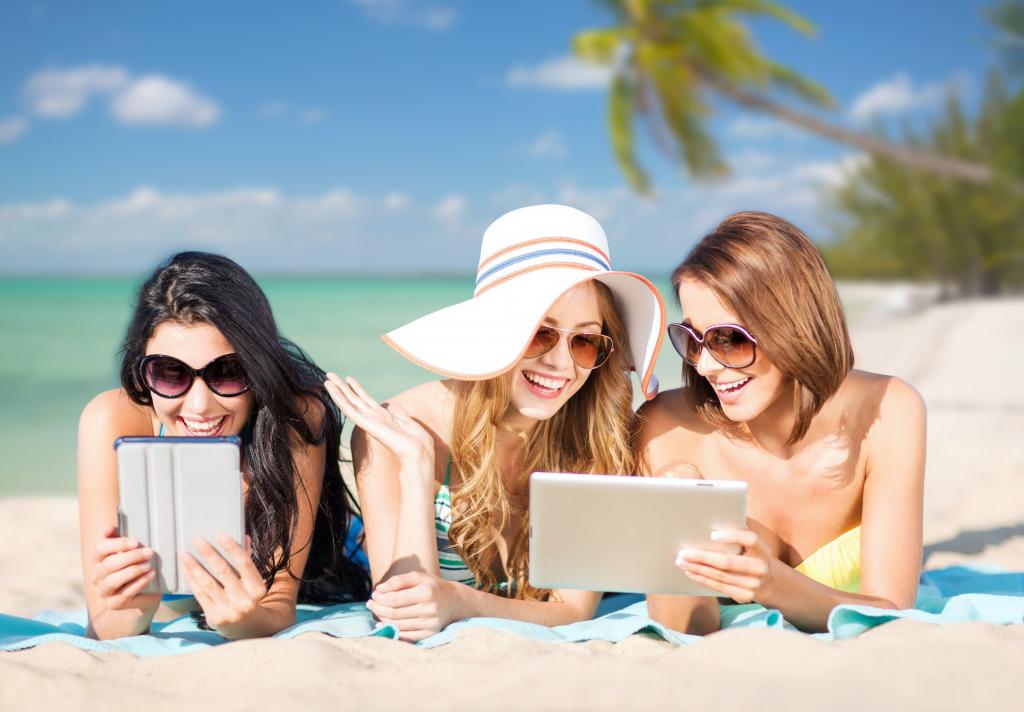 三个微笑女孩在手中的平板电脑在沙滩上晒日光浴