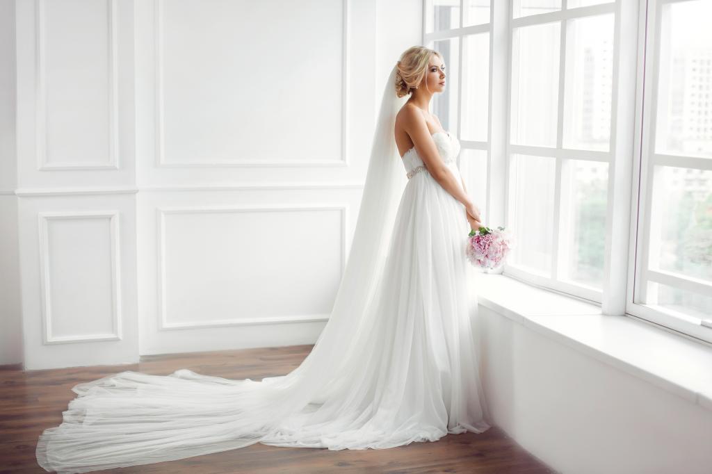 美丽的女孩在一束白色的婚纱礼服站在窗户旁边