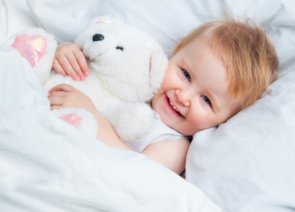 一个微笑的孩子躺在床上，一个白色的泰迪熊